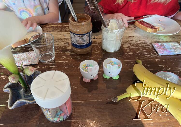 Image shows the girls decorating their toast with the jar of peanut butter and cream cheese between them. The bunnies sit facing the camera by the bananas and main sprinkle container. 