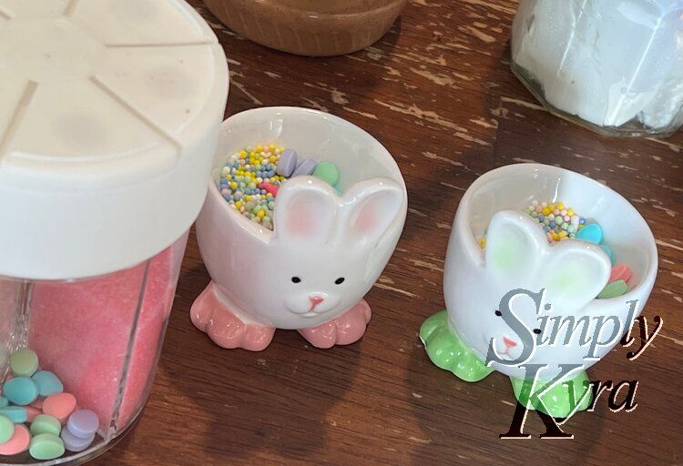 Image shows two bunny shaped egg cups next to a large divided container of sprinkles. 