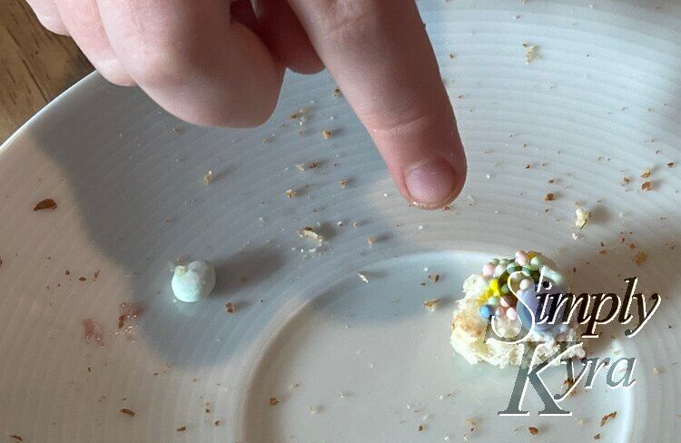 Image shows a small piece of bread covered in sprinkles next to a blue sprinkle to the side. A finger is pointing down at the bread so it's not missed in the photo. 