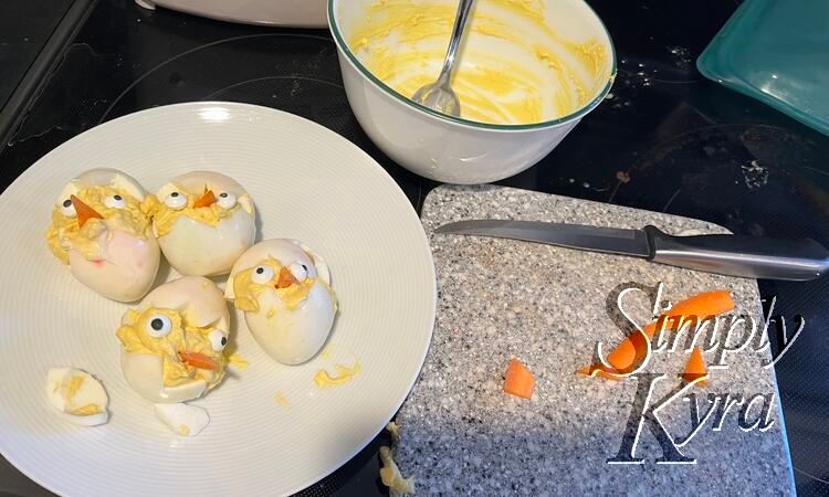 Image shows a plate with four finished chicks and two pieces of egg white. To the right sits a small marbled cutting board with a knife and carrot bits. Behind it all sits a dirty white bowl with the yolk mixture remnants. 