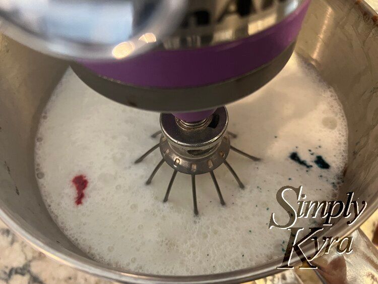 Image shows the bowl of a stand mixer looking down with the whisk down and turned off. There's red dye to the left and blue to the right. 