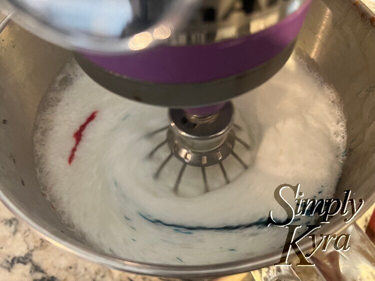 Image shows the bowl of a stand mixer looking down with the whisk down, turned on, and blurred. The red has streaked about an inch and the blue has been brought around and going up. Each droplet is now a dahs further to the left. 
