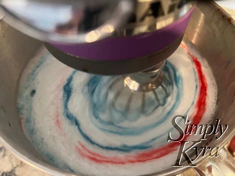 Image shows the bowl of a stand mixer looking down with the whisk down, turned on, and blurred. Both colors have been streaked around and there might be a faint lavender hue formed.