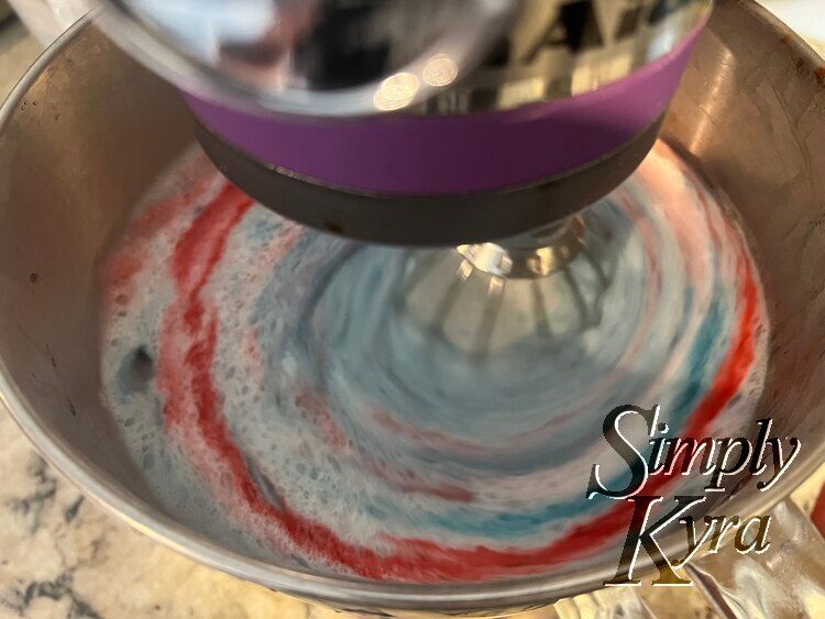 Image shows the bowl of a stand mixer looking down with the whisk down, turned on, and blurred. Way more bubbles and all angel food cake is red, blue, or off white. 