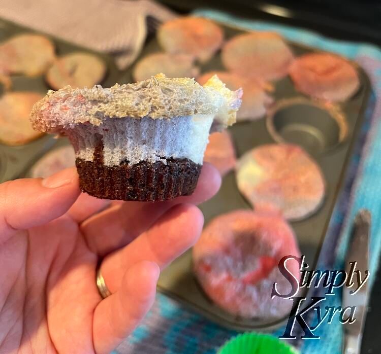 Image shows a whole cupcake without liner being held up in front of the blurred pans. 