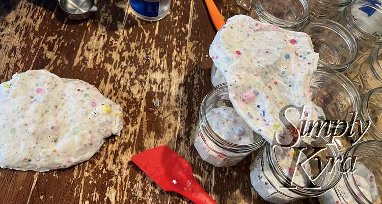 Image shows four jam jars partially filled with the slime with another section to the left loose and undivided on the table. Over the jars sits one section pulled apart to better highlight the items inside it. 