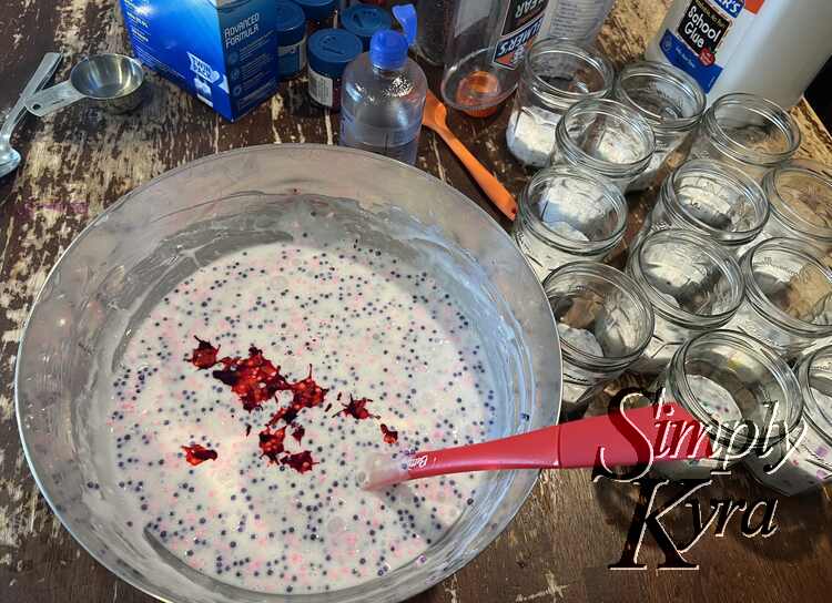 The bowl is filled with white slime speckled with pink and purple foam balls. There's red splotches on top. To the right sits the jam jars with white multicolor spotted slime. 