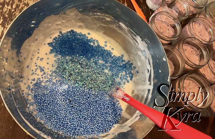 Image shows blue foam balls, glitter, and plastic gems sitting on top of the white glue in the bowl. A red spatula sits within and the jars to the right are topped off with purple slime. 