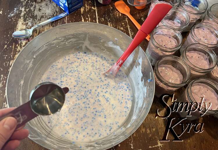 Image shows the white bowl with blue spotted white slime. A measuring cup is pouring clear liquid into the bowl and the jam jars sit to the side with the lavender slime in them. 