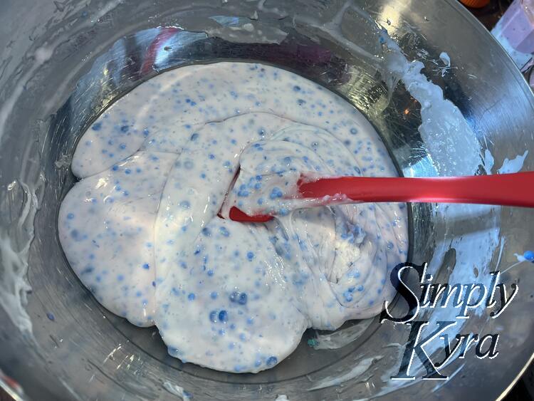 Image shows a closeup of the metal bowl with white blue specked slime and a red spatula inside. There's clear separated liquid around the perimeter.