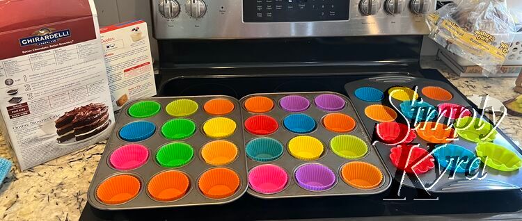 Image shows the box mix brownies and angel food cake to the left (showing the directions) and three cupcake pans with colorful silicon cupcake liners laid out side by side on the stovetop. 