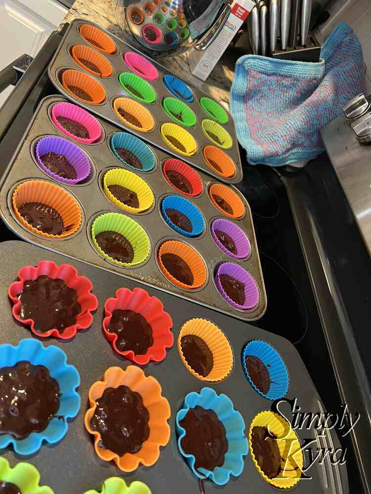 Image shows the same stove but this time from the right. All three dozen liners are a third filled with brownie mix. The metal stand mixer bowl sits to the side (background) reflecting the image back at us. 