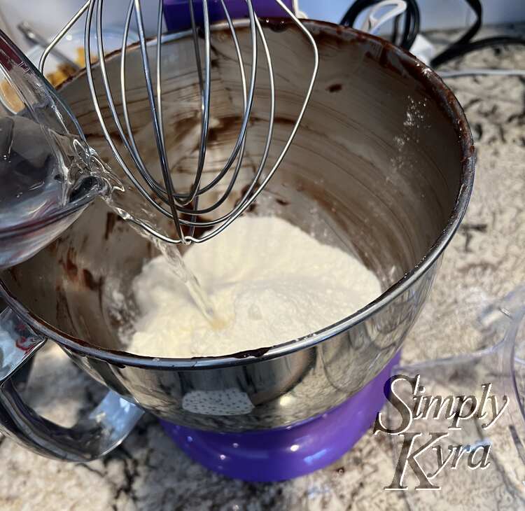 Image shows the brownie streaked stand mixer bowl with a pile of white at the bottom and water being poured in. The whisk is above poised to be lowered.