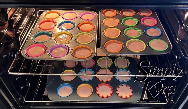 Three cupcake pans are in the oven. The top layer has two dozen side by side while the bottom shelf has a larger pan. 