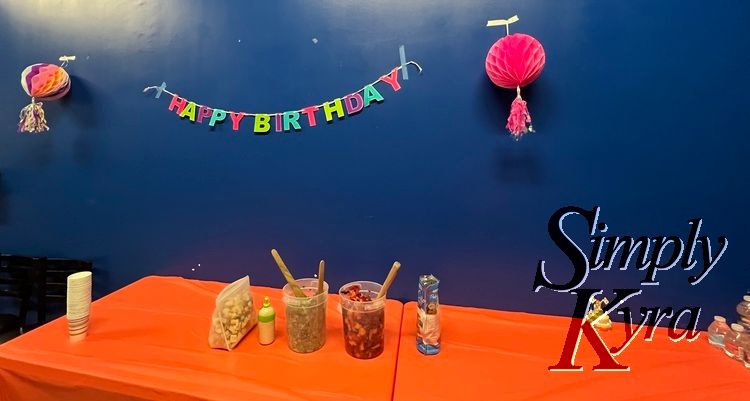Image shows a bright orange table in front of a deep blue wall. There's a glittery Happy Birthday sign above with two tasseled paper globes from last year's party. I brought our LEGO decoration although I don't think the kids noticed it. 