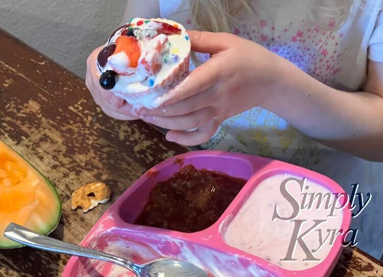 Image shows a pink divided plate with fruit salad leftovers and yogurt. Zoey holds the cupcake above it with blue sprinkles, strawberries, blueberries, red sauce, and whip cream. 