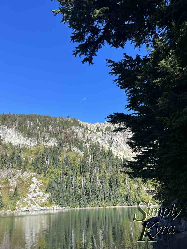 Image shows an airplane trail in the sky with the fire lookout right below it. The trees on the hill are reflecting in the water.