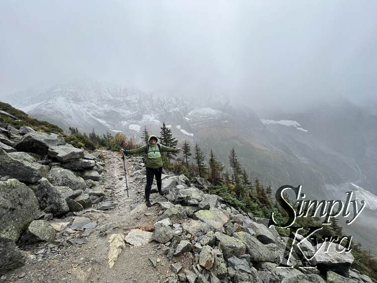 I stand with my arms outstretched along the rocky path with smaller trees along the edge of the hill. Foggy mountains lie behind in the background.