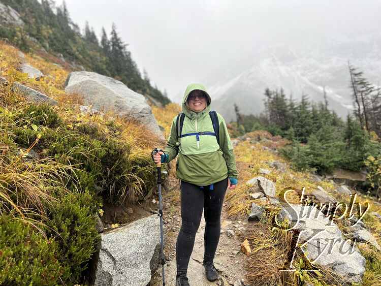 I stand smiling on the dirt path surrounded by yellowed plants and large boulders. The background of foggy mountains and trees is blurred. 