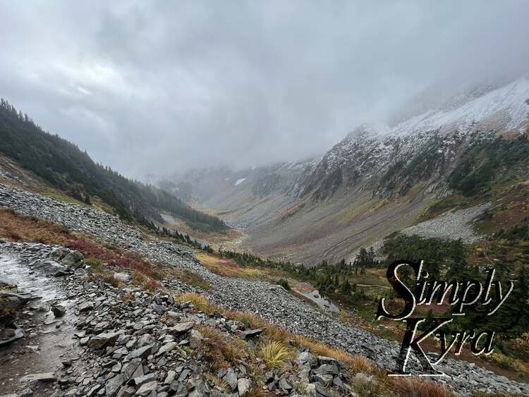 Image shows a colorful vista of yellow, red, and grey with hills of trees, rock, and snow. A path cuts through on the left side.