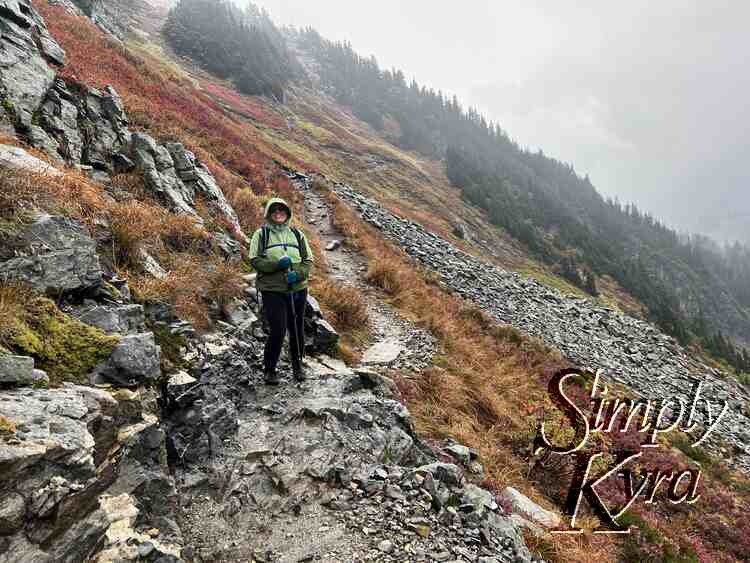 I stand in the center of a rocky path cutting through a field of yellow, red, and green grass and rocks. Trees and talus fields sit behind. 