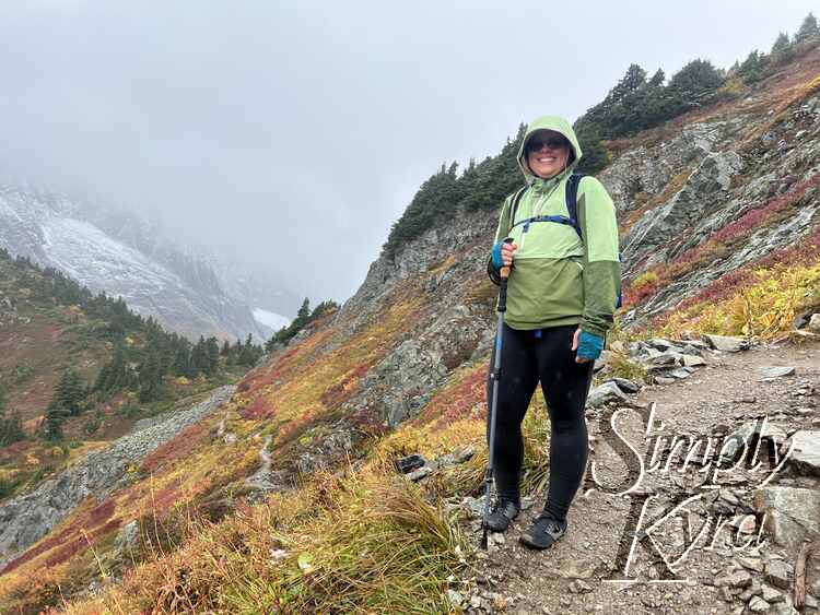 Image shows me at the foreground on the dirt path with the colorful hill falling away to the left and climbing on the right. In the background s another small colorful hill and talus to the left and grey of snow, cloud, and mountain in the background.