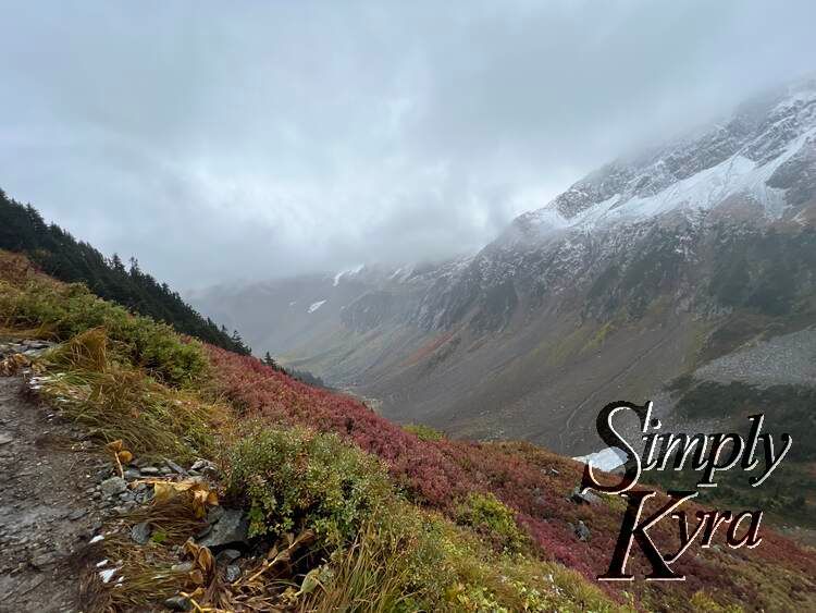 Image shows a yellow, red, and green hill rising to the left with the grey-ish mountain to the right and clouds roiling in between. 