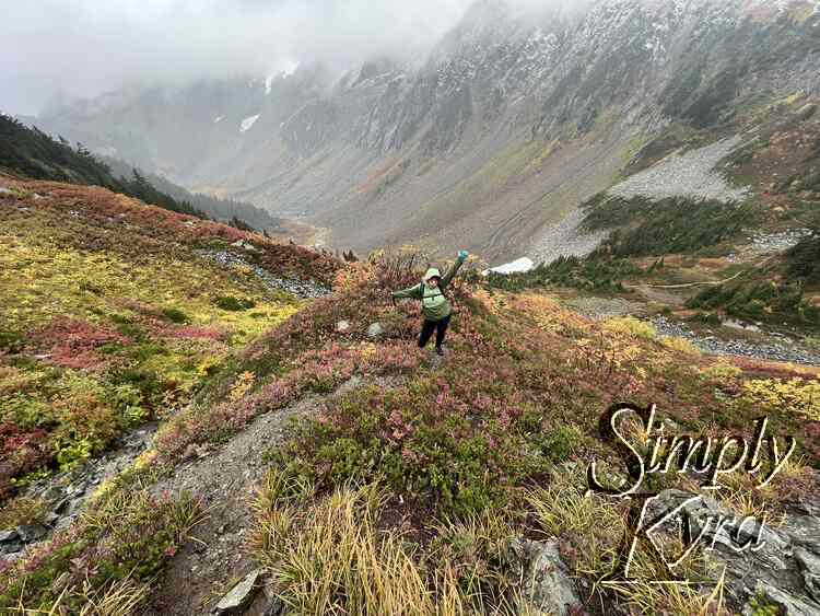 I stand with my arms outstretched and tongue out on the pathway surrounded by colorful hills in red, yellow, and green. In the background is the base of a grey background. 