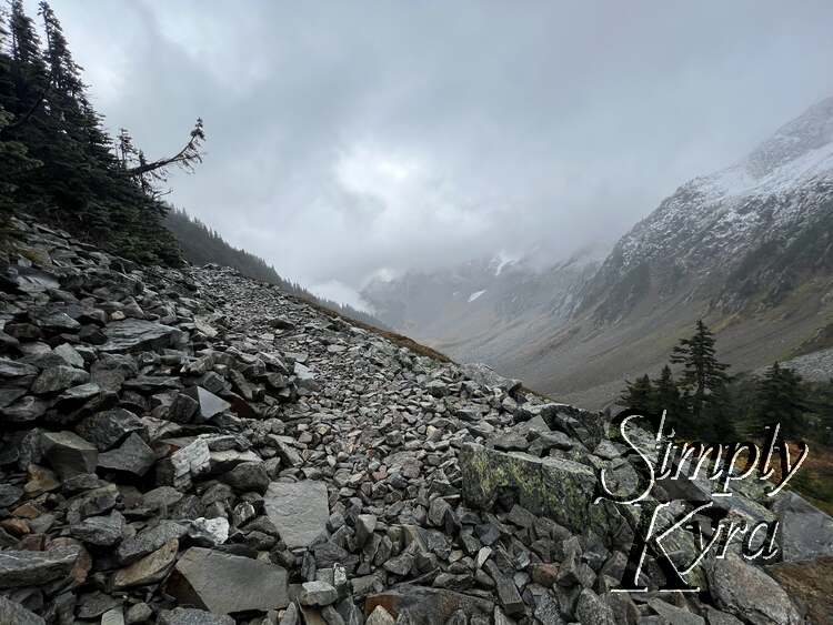 A hill of rock lays along the left and center of the image with trees to the left and right. In the background sits the snow capped mountain and clouds. 