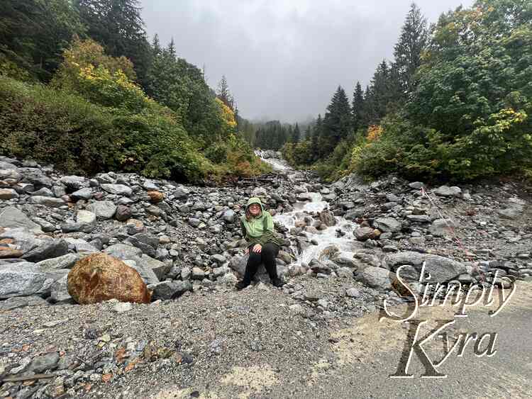 I sit on a rock in front of the creek. The waterfall is behind me with the green trees to either side in the background. In the foreground to the right lays the road. 