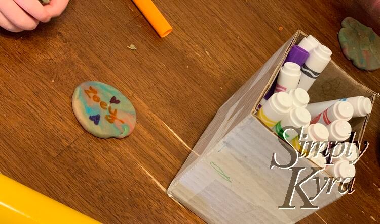 Image shows a simple marbled disc of playdough saying Zoey with two hearts above and below beside a box of washable markers.