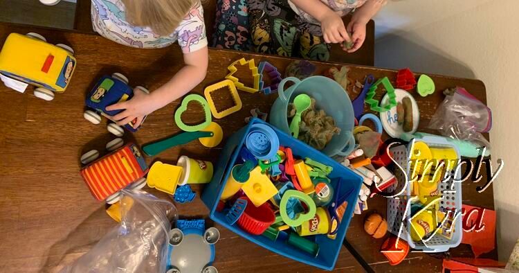 Image shows a table strewn with playdough toys, playdough, and toy cars along with two kids.