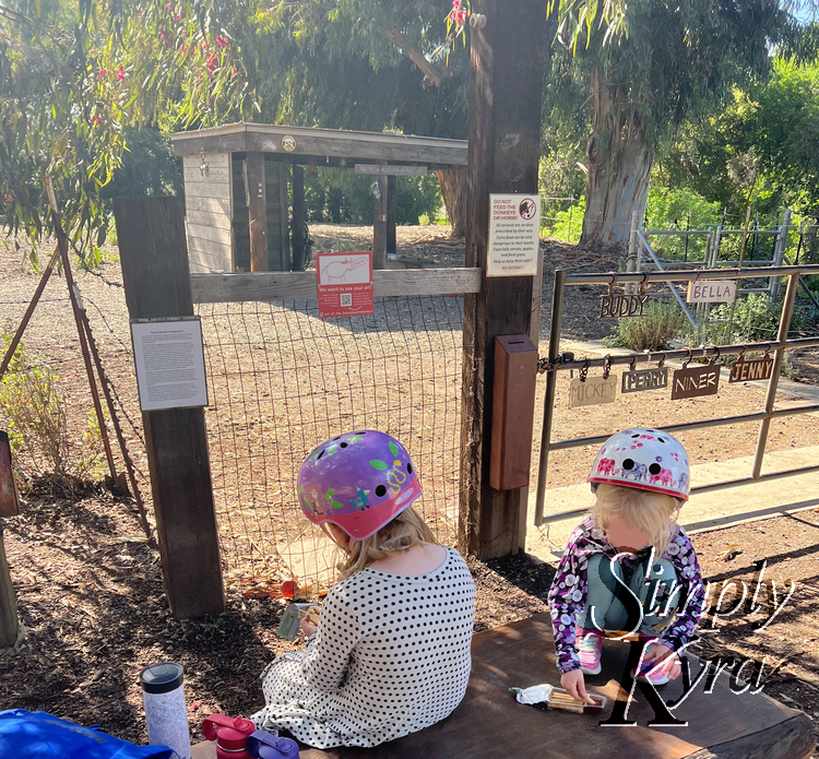 Image shows the girls sitting on the bench in front of the labelled gate eating a snack.