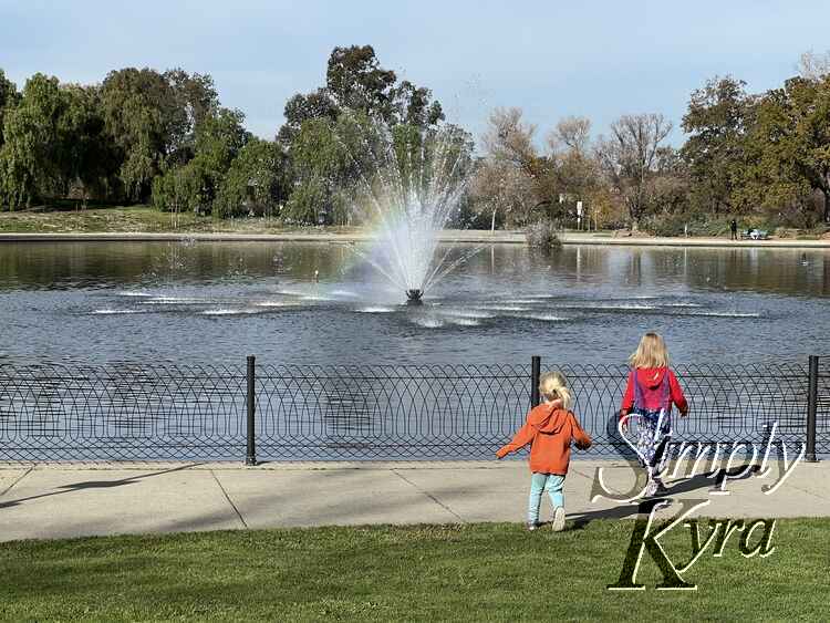 Kids approaching the fence surrounding the lake with a fountain and rainbow beam.