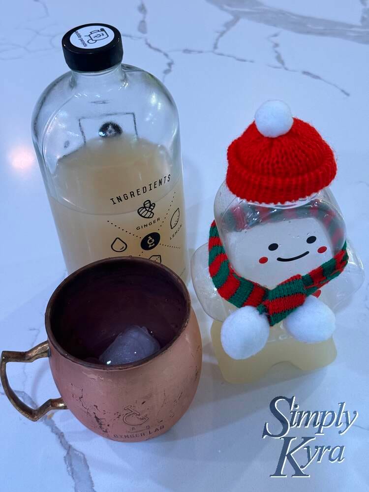 Image shows a partially filled festive ginger beer bottle standing next to an ice added Ginger Lab mule glass with a two thirds full growler behind it. 