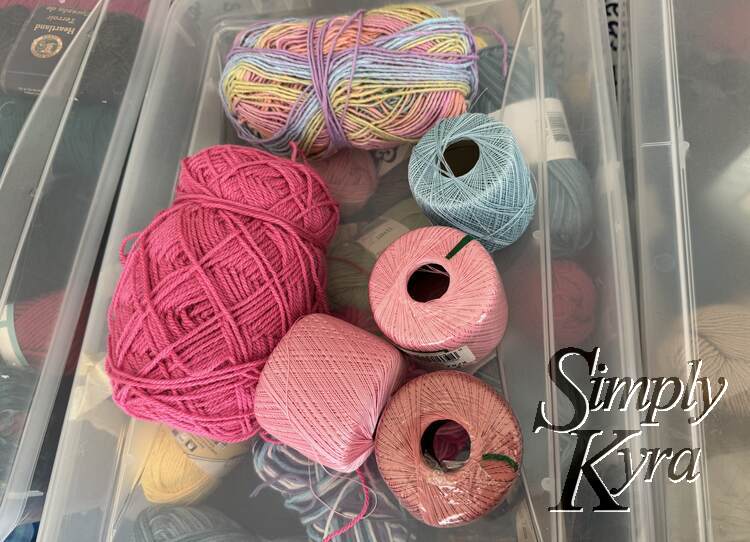 Two unlabelled balls of yarn and four rolls of crochet thread on top of a clear bin full of yarn.