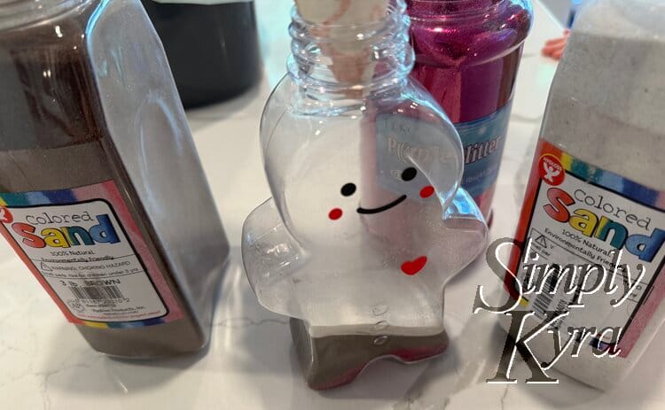 Image shows the ginger bread bottle with no lid and the paper funnel in. It's surrounded by the sand and glitter container and has purple, brown, and white layers near the bottom.