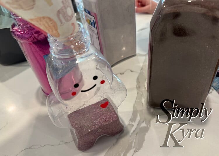 Image shows the ginger bread bottle with no lid and the paper funnel in. It's surrounded by the sand and glitter container and has a purple tinged brown sand at the bottom. 