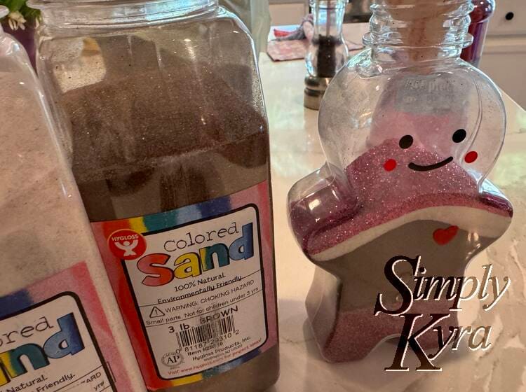 Image shows the ginger bread bottle with no lid and the paper funnel in. It's surrounded by the sand and glitter container and has a purple tinged brown sand at the bottom then a large amount of brown and smaller layers of white and purple.