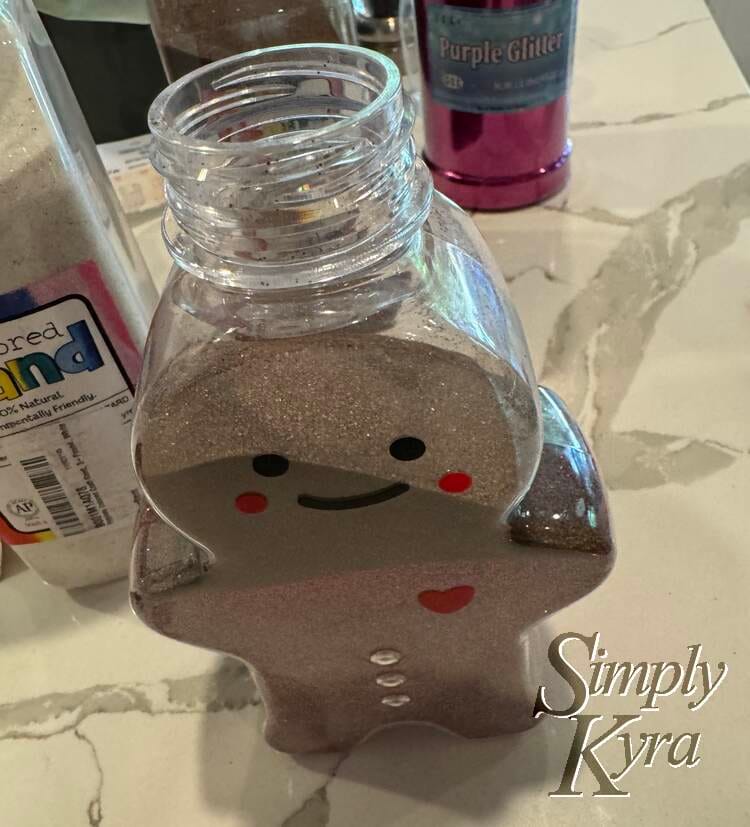 Image shows the ginger bread bottle with no lid and the paper funnel in. It's surrounded by the sand and glitter container and has a purple tinged brown sand at the bottom with brown covering half the face. 