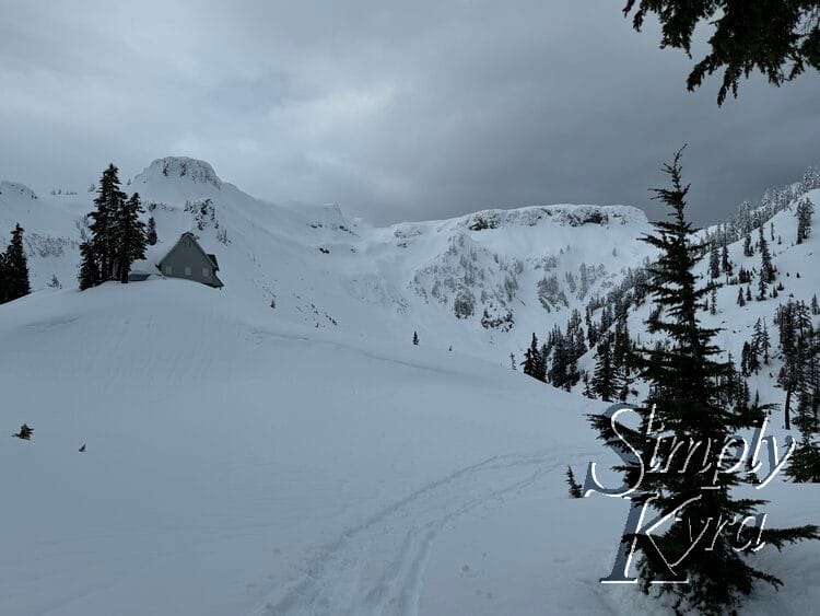 A trail goes in front with trees to the left, the snow covered building to the right on a hill, and mountains in the background. 