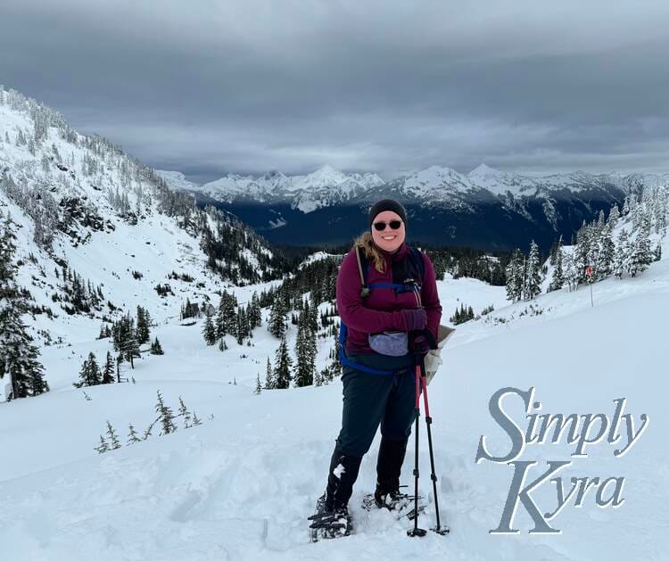 Image shows me in snowshoes with my poles grasped in one hand standing smiling with snow covered hills and mountains in the background. 