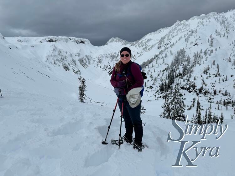 Image shows me in snowshoes with my poles grasped in one hand standing smiling with snow covered hills and a ridgeline.