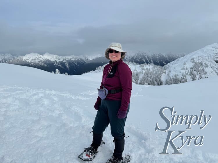 Image shows me in snowshoes with my poles grasped in one hand standing smiling with snow covered hills and mountains in the background. 