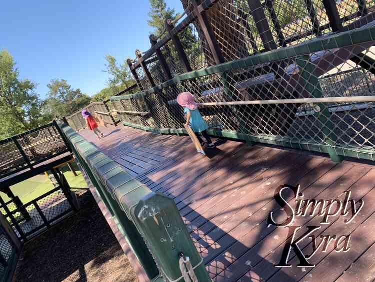 Image shows the girls running along a wide wooden platform towards the hill in the background. Both are carrying a piece of cardboard to slide in more ways. 