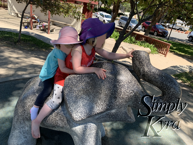 Image shows Zoey hugging Ada's waist as both girls sit on a concrete elephant. 
