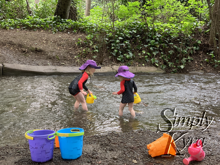 Image shows the girls side by side in matching sunhats and swimsuits each holding a plastic yellow watering can with other buckets on the side of the creek. 