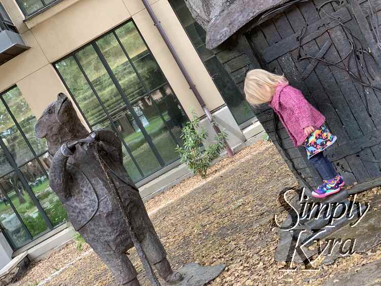 Image shows Zoey posed by the metal door in the tree while a statue watches on. 