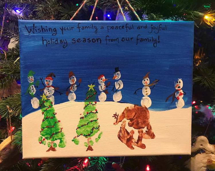 Image shows a canvas hanging by multicolored yarn in front of a Christmas tree. On the canvas it says "Wishing your family a peaceful and joyful holiday season from our family!". Below the words are fingerprint snowmen, two footprint decorated trees, and a handprint reindeer on snow. 