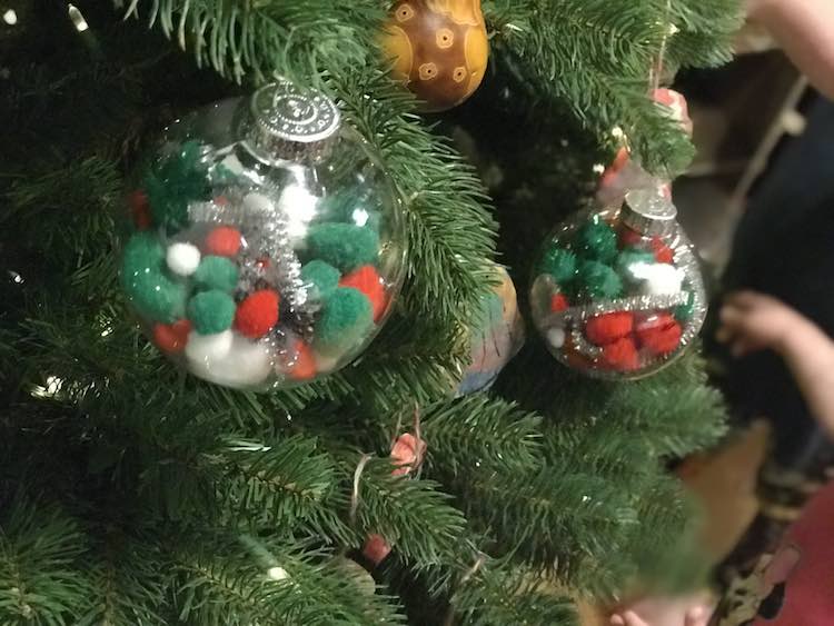 Image shows the pompom and pipe-cleaner filled plastic orbs on the Christmas treed with a kid blurred out in the background playing with their kitchen set. 